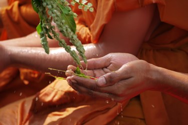 Close-up of a Buddhist monk in saffron robes performing a traditional water blessing ceremony, gently pouring water over hands during a spiritual ritual in Southeast Asia.