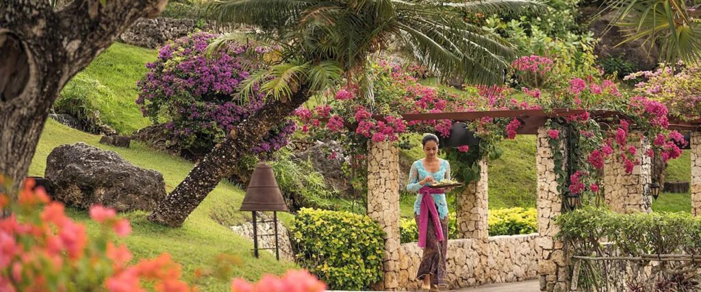 Woman in traditional Balinese attire walks through a flower-lined garden at Four Seasons Resort Bali at Jimbaran Bay, a romantic honeymoon destination surrounded by lush tropical greenery.