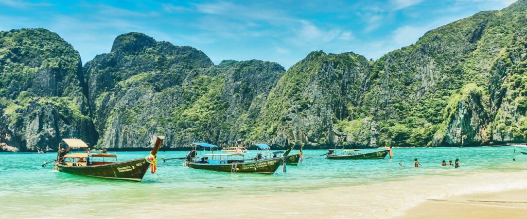 Beach scene in Krabi, Thailand, with traditional long-tail boats anchored in turquoise water, limestone cliffs rising in the background, and swimmers enjoying the shallow shoreline.