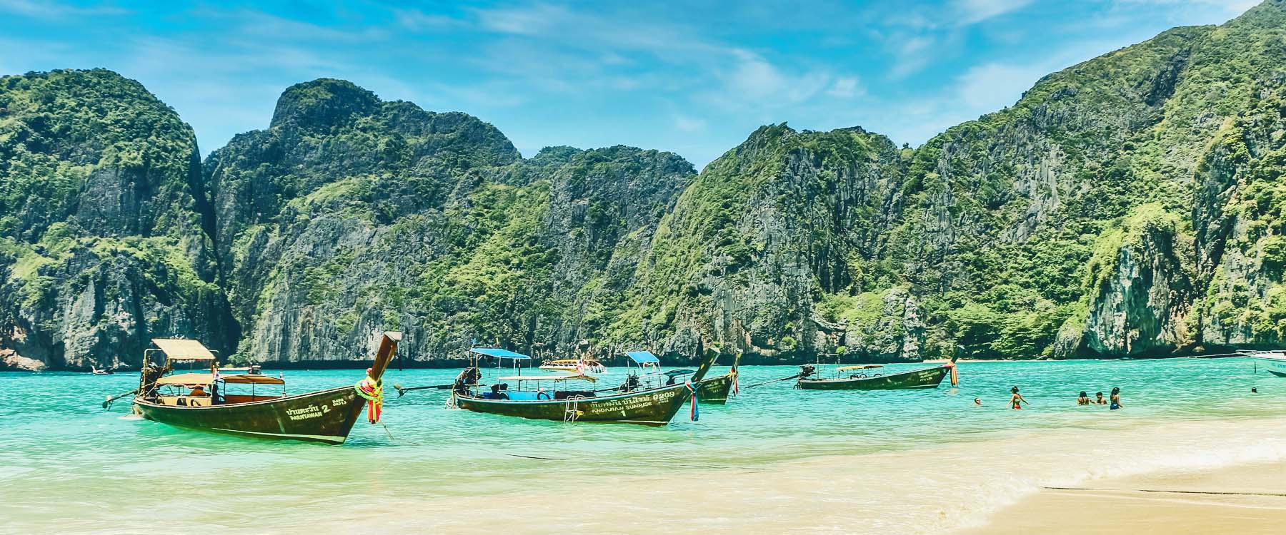 Beach scene in Krabi, Thailand, with traditional long-tail boats anchored in turquoise water, limestone cliffs rising in the background, and swimmers enjoying the shallow shoreline.