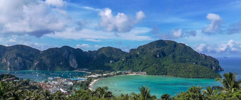 Panoramic view of Phuket, Thailand, featuring a turquoise bay with boats anchored near a coastal village, framed by lush green mountains under a partly cloudy sky.