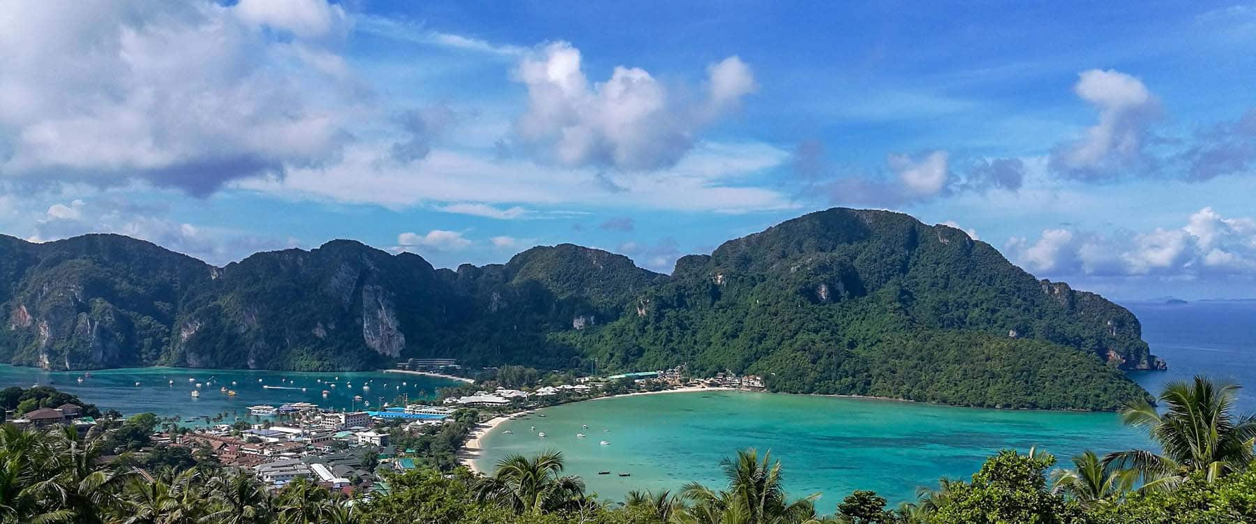 Panoramic view of Phuket, Thailand, featuring a turquoise bay with boats anchored near a coastal village, framed by lush green mountains under a partly cloudy sky.