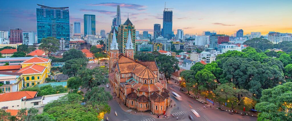 Aerial view of Ho Chi Minh City featuring Notre-Dame Cathedral Basilica in the foreground, surrounded by tree-lined streets and colonial buildings, with modern skyscrapers rising in the background at dusk.