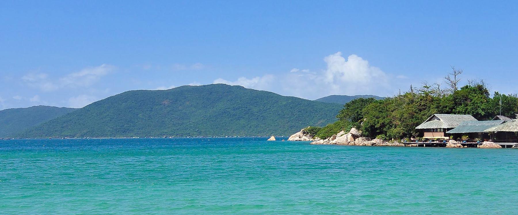 Coastal view of a quiet beach in Vietnam with clear turquoise water, a small wooden pavilion by the shoreline, and lush green hills rising across the bay under a bright blue sky.