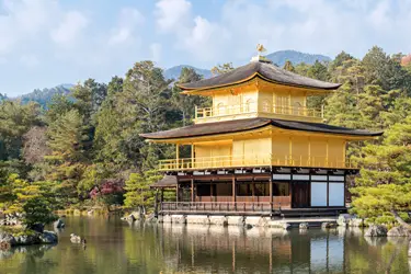 Kinkaku-ji (Golden Pavilion) in Kyoto reflected on a tranquil pond, surrounded by forested hills and traditional Japanese gardens.