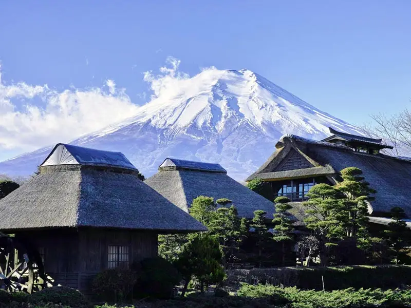 Mount Fuji towering above traditional thatched-roof houses in a historic village near Lake Kawaguchi.