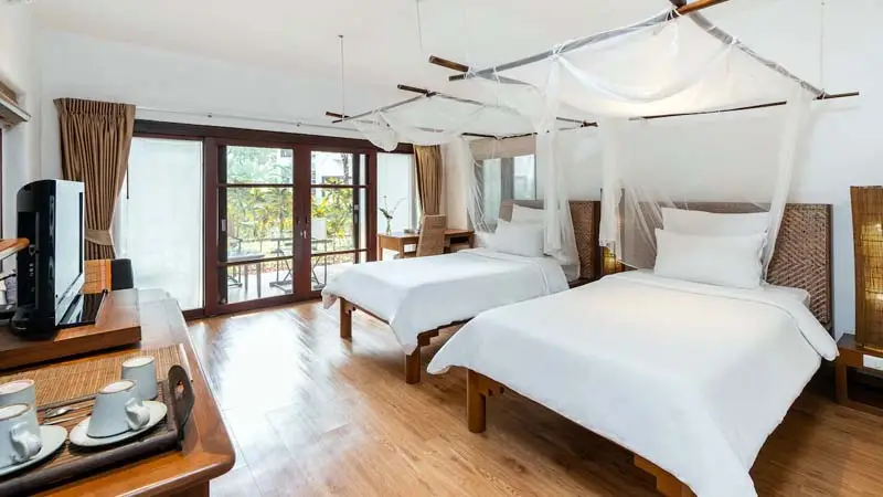 Guest room at Chiang Rai Legend Boutique Hotel, featuring twin beds with canopy netting, natural wood furnishings, and garden-facing windows in a serene riverside setting.