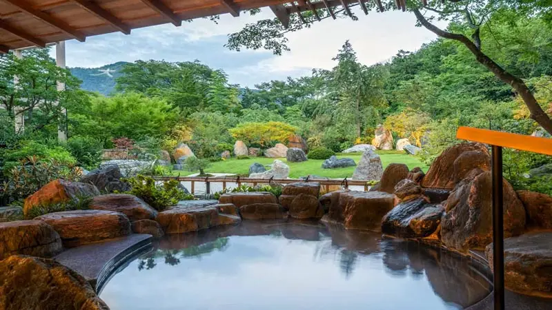 Open-air onsen bath at Gora Kadan Ryokan in Hakone, overlooking a serene Japanese garden with rocks, greenery, and forested hills.