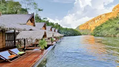 Thatched-roof floating lodge along the River Kwai in Kanchanaburi, Thailand, with wooden decks, riverfront loungers, and lush jungle scenery.