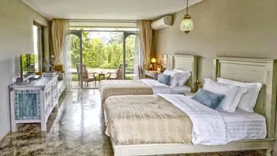 Elegant guest room at The Reverie Siam Mae Hong Son, featuring vintage-inspired décor, twin beds, and large windows overlooking lush gardens in a serene riverside setting.