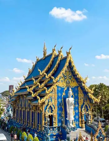 Wat Rong Suea Ten (Blue Temple) in Chiang Rai, Thailand, featuring vivid blue and gold architecture with ornate carvings and a white Buddha statue under a clear sky.