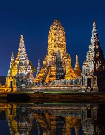 Wat Chaiwatthanaram temple in Ayutthaya, Thailand illuminated at night, featuring towering prangs reflected in still water within the historic ruins complex.