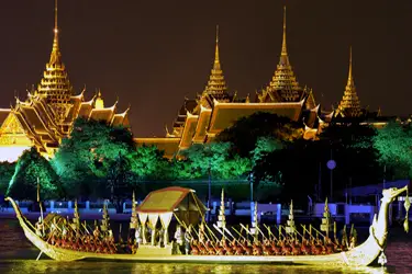 Royal Dragon boat illuminated at night on the Chao Phraya River in Bangkok, Thailand, with the Grand Palace’s golden spires glowing in the background.