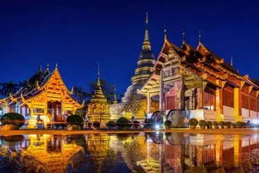 Wat Phra Singh temple complex in Chiang Mai, Thailand at night, showcasing golden stupas, ornate Lanna-style buildings, and reflections shimmering on a tranquil water surface.