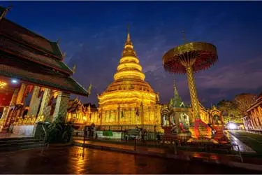 Illuminated Buddhist temple in Chiang Mai, Thailand at night, featuring a golden chedi, ornate Lanna architecture, and ceremonial umbrellas glowing against the evening sky.