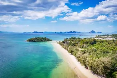 Aerial view of Krabi’s coastline in Thailand, showcasing a sweeping sandy beach, turquoise Andaman Sea waters, lush greenery, and limestone islands on the horizon.