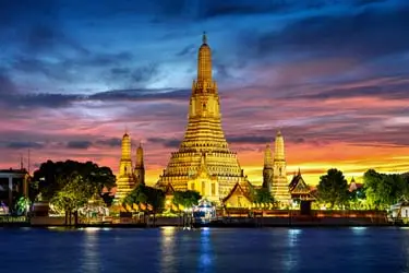 Wat Arun (Temple of Dawn) in Bangkok, Thailand illuminated at sunset, with its ornate spires reflecting across the Chao Phraya River.