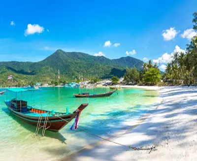 Long-tail boats anchored along a white-sand beach on Koh Phi Phi, Thailand, with crystal-clear turquoise water, lush tropical hills, and a bright blue sky.
