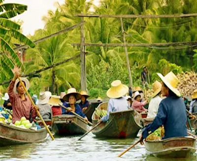 Damnoen Saduak Floating Market in Thailand, with local vendors paddling wooden boats along narrow canals amid lush palm trees and traditional riverside scenery.