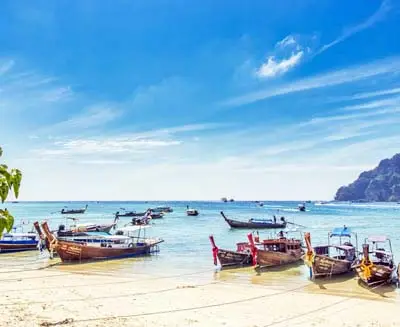 Long-tail boats lined up along the sandy shore of Phi Phi Islands, Thailand, with turquoise waters, limestone cliffs, and a bright tropical sky.