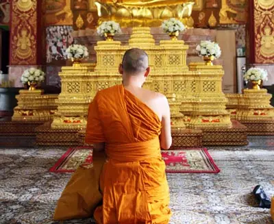 Buddhist monk kneeling in prayer inside a traditional Thai temple, facing a golden altar adorned with ornate carvings and floral offerings.