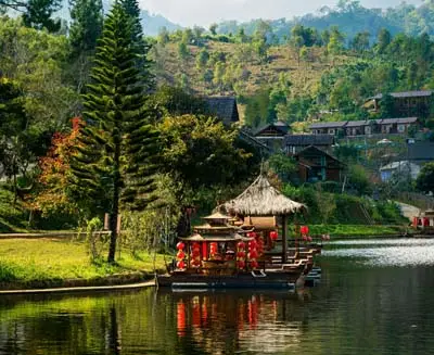 Traditional lakeside pavilion along the Mae Ping River in northern Thailand, reflected in calm water and framed by forested hills and hillside village homes.