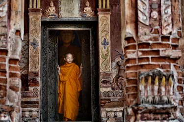 Buddhist monk in saffron robes standing in an ancient Angkor temple doorway in Cambodia, framed by weathered stone carvings.