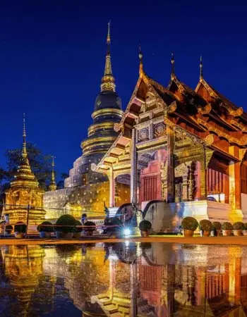 Wat Phra Singh temple complex in Chiang Mai, Thailand at night, featuring golden stupas, ornate Lanna architecture, and reflections shimmering in a tranquil water feature.