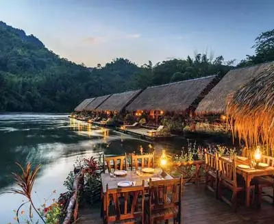 Riverside dining at a floating lodge along the River Kwai in Kanchanaburi, Thailand, with lantern-lit tables, thatched-roof bungalows, and tranquil jungle surroundings at dusk.