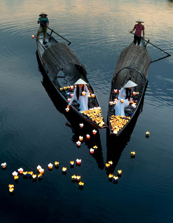 Women releasing floating lanterns from traditional wooden boats on the Thu Bon River in Hoi An, Vietnam, capturing a serene cultural ritual and romantic riverside atmosphere.