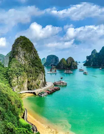Aerial view of Ha Long Bay, Vietnam, showcasing emerald waters, limestone karst islands, and traditional boats along a secluded sandy shoreline under a bright blue sky.