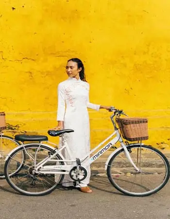 Woman in a traditional white áo dài standing beside a bicycle against a vibrant yellow wall in Hoi An, Vietnam, reflecting the town’s timeless charm and cultural elegance.