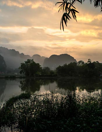Sunset over limestone karst mountains reflected in a tranquil river in Ninh Binh, Vietnam, showcasing lush wetlands and the region’s iconic dramatic landscape.