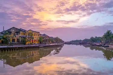 Hoi An ancient town riverside at sunset with lantern-lit heritage buildings, calm canal reflections, and traditional boats along the Thu Bon River in central Vietnam