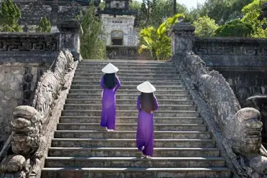 Two women in traditional áo dà i and conical hats ascending stone steps at an ancient imperial site in Vietnam, framed by historic architecture and lush greenery.