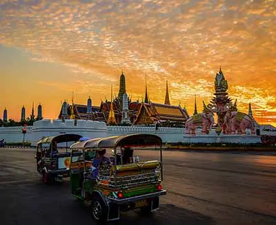 Tuk-tuks driving past the Grand Palace and Wat Phra Kaew at sunset in Bangkok, Thailand, with golden temple spires glowing under a dramatic orange sky.