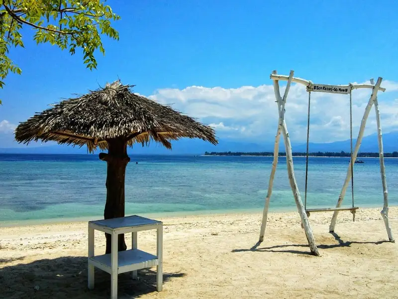 Gili Air island beachfront with traditional wooden swing, shaded thatched umbrella, and calm turquoise waters along the Lombok Strait