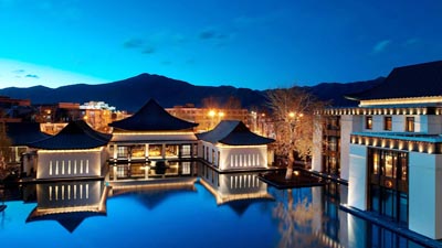 The St. Regis Lhasa Resort at dusk, featuring Tibetan-inspired architecture reflected in a tranquil pool, with mountain silhouettes and city lights in the background.