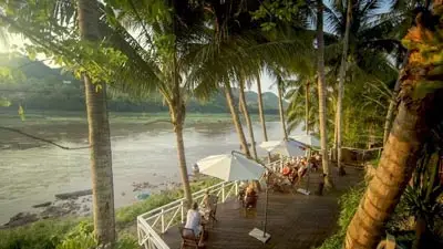 Belle Rive Boutique Hotel terrace in Luang Prabang overlooking the Mekong River, with palm trees, wooden deck seating, and sunset river views in Laos.