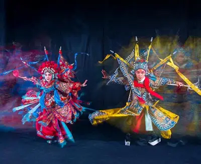 Traditional Chinese opera performers in elaborate costumes and face makeup, captured in dynamic motion during a colorful stage performance.