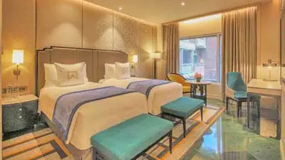 Elegant guest room at ITC Rajputana, Jaipur, featuring twin beds with upholstered headboards, teal benches and chairs, warm ambient lighting, and a large window with city views.