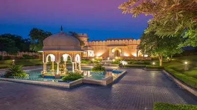 Evening view of The Oberoi Rajvilas, Jaipur, featuring a domed pavilion over a reflective pool, landscaped gardens, and softly lit palace-style architecture.