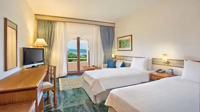 Bright guest room at Trident Jaipur featuring twin beds, a work desk, and a private balcony with views of greenery and surrounding hills.