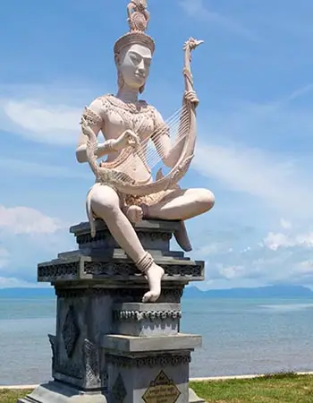 Statue of Yeay Mao (Grandmother Mao) by the sea in Kep, Cambodia, depicting the revered local spirit holding a traditional instrument against a coastal backdrop.