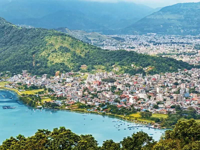 Aerial view of Pokhara, Nepal with Phewa Lake in the foreground, dense city neighborhoods along the shoreline, and green Himalayan foothills rising in the background.
