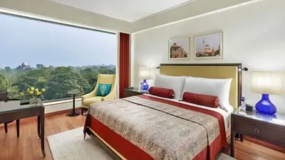 Modern guest room at The Oberoi, New Delhi, featuring a king bed with red accents, large picture window overlooking greenery, and a stylish seating area.
