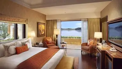 Luxury room at The Leela Palace Udaipur featuring a king bed with warm-toned decor, elegant seating area, and a balcony overlooking Lake Pichola and surrounding hills.