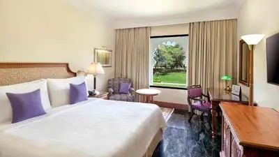Guest room at Trident Udaipur featuring a king bed with purple accents, classic furnishings, and a large window overlooking lush gardens.