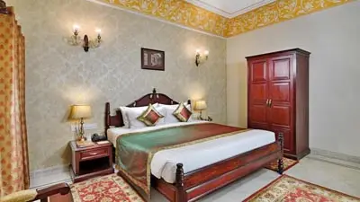 Classic heritage-style room at BrijRama Palace, Varanasi, featuring a wooden bed with ornate headboard, patterned textiles, antique furnishings, and warm traditional decor.