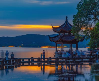 West Lake at dusk in Hangzhou with a traditional Chinese pavilion reflected on calm water and surrounding hills.